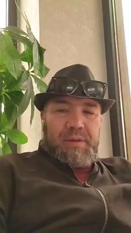 The video shows a close-up, head-and-shoulders shot of a man wearing a hat and sunglasses. He has a beard and is looking directly at the camera. The background appears to be a wall with a textured pattern and a large green plant to the left. The man's mouth is slightly open, as if he is speaking or about to speak. His expression is neutral. The lighting suggests it is daytime, possibly indoors.
