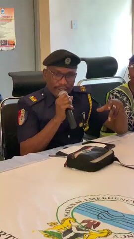 A man wearing a dark blue uniform with a beret, glasses, and a microphone is speaking. He is sitting at a table covered with a white cloth that has a logo on it. He is gesturing with his right hand. In front of him on the table is a small black bag and some papers. Behind him, there are chairs and a poster on the wall. Another person, only partially visible, is sitting to his right. The man appears to be giving a presentation or speech.