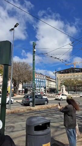 The video shows a street scene in a European city, possibly Lisbon, on a sunny day with scattered clouds.  Several people are waiting at a crosswalk, some taking pictures of the buildings.  The architecture is characterized by multi-story buildings with colorful facades, including shades of yellow and blue, and orange tiled roofs.  There are tram tracks on the street, and a yellow tram is seen passing by. Cars are also driving on the street.  Overhead are numerous power lines for the tram system. Streetlights and utility poles are visible throughout the scene, along with a trash can. Construction activity, indicated by a crane, is present in the background.  The overall impression is of a bustling urban environment with a mix of architectural styles.