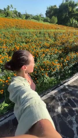 The video shows a woman taking a selfie in front of a large field of orange marigolds. The woman is standing on a gray brick structure. She has her hair pulled back in a bun and is wearing a light green shirt. She is smiling and holding the camera out in front of her. The field of marigolds stretches up a hillside. The sky is blue with a few white clouds. Trees and a couple of buildings can be seen in the background.