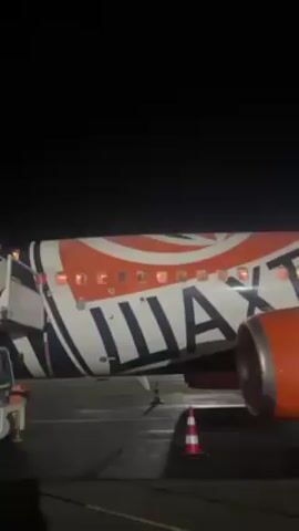 The video shows the side of a stationary airplane at night, likely on a tarmac. The plane is painted with a distinctive orange, white, and black color scheme, and the word "Шахтар" (Shakhtar) is prominently displayed. Interior lights are visible through the windows of the plane. An orange jet engine is attached to the wing, and a red and white traffic cone stands on the ground near the plane. The background is dark and lacks specific detail.