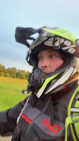 A person wearing a helmet and safety gear is riding a dirt bike along a grassy path. The camera angle shifts between a view of the rider's face and a first-person perspective looking down at the bike and the path ahead. The environment consists of green grass, trees, and a cloudy sky. The overall impression is one of an off-road recreational activity.
