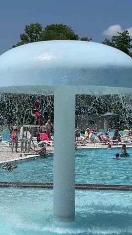 The video shows a busy swimming pool on a sunny day. In the foreground, a mushroom-shaped water feature sprays water into the pool, creating a curtain of water. In the background, people of various ages are swimming, wading, and relaxing on lounge chairs. There are trees and a blue sky with white clouds. The pool has a tiled edge with a blue and white pattern.