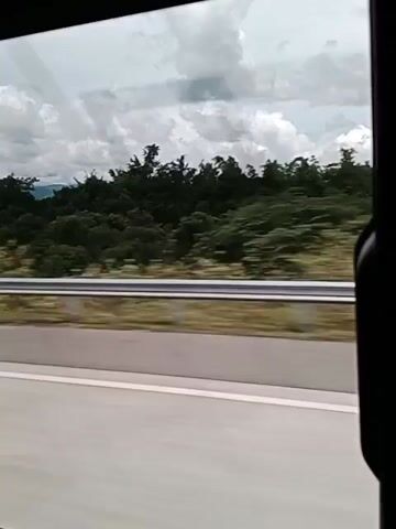 This is a first-person perspective video captured from a vehicle, likely a car or bus, traveling on a highway. The camera is pointed out the side window. The foreground shows a paved road with a white line. Beyond the road, there's a silver guardrail, and behind that, a dense landscape of green trees and foliage. In the distance, hills or mountains are visible, covered in trees, under a cloudy sky. The video is short, showing the scenery passing by. There are no people or discernible actions visible within the frame. The motion blur indicates the vehicle is in motion.