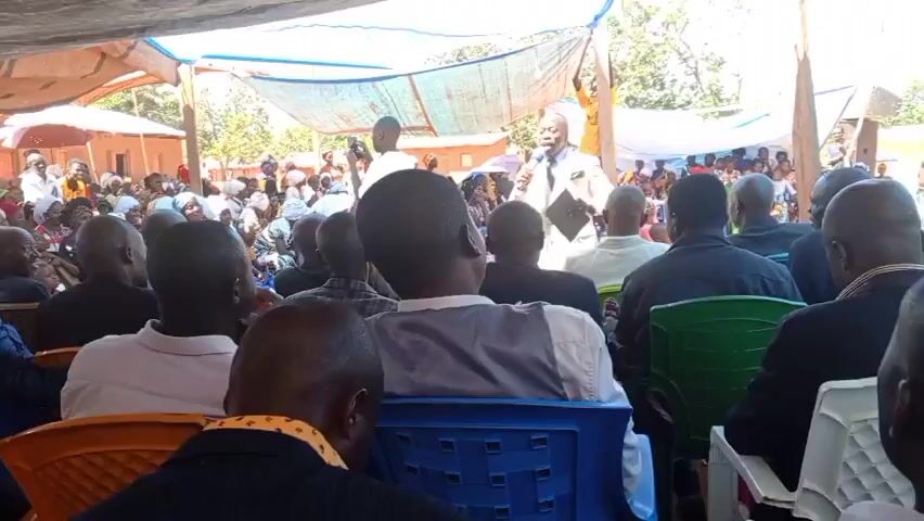 A man in a white suit is speaking into a microphone in front of a crowd of people seated under a canopy. He is holding a black book in his other hand. The crowd appears to be of African descent and is listening attentively. There is a person recording the event with a cell phone. The scene appears to be outdoors, with trees and buildings visible in the background.