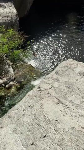 A first-person perspective video begins with a view of a natural swimming area with dark water and sunlight reflecting off its surface. A person is partially visible on the left, appearing to be in the water or about to enter it, with a splash indicating movement. The edge of a large, light-colored rock occupies the foreground.

As the video progresses, the camera pans slightly to reveal more of the surrounding environment. Several people are scattered on rocks and along the bank of the water. Some are sitting, while others appear to be wading or swimming. In the background, a multi-story building with red umbrellas on an outdoor seating area is visible, nestled among lush green trees. A white inflatable ring floats near the rocky bank. The overall scene depicts a popular outdoor recreational spot on a sunny day.