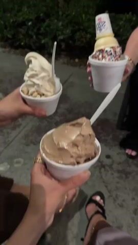Three individuals hold cups of ice cream. The flavors are vanilla with nuts, vanilla with a cone and sprinkles, and chocolate. They stand outdoors at night with green foliage visible in the background. Two people wear open-toe sandals. The background includes pavement and an unseen structure.