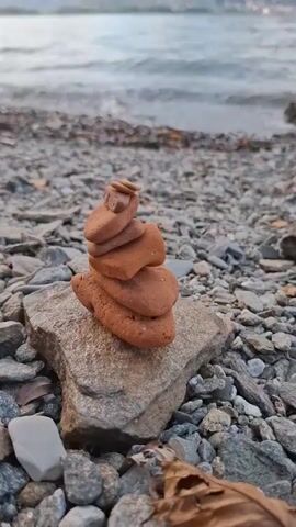 The video shows a cairn, or stack of balanced stones, on a rocky shore. The stones are reddish-brown and appear to be smooth. The cairn is resting on a larger, flat rock. In the background, a body of water, likely a lake or sea, is visible. Waves are gently lapping at the shore. Further in the distance, hills or mountains rise, covered in trees and some buildings. The sky is cloudy. The perspective is low, focusing on the cairn and the immediate surroundings. The video seems to be a close-up shot, emphasizing the texture of the stones and the pebbles on the shore.