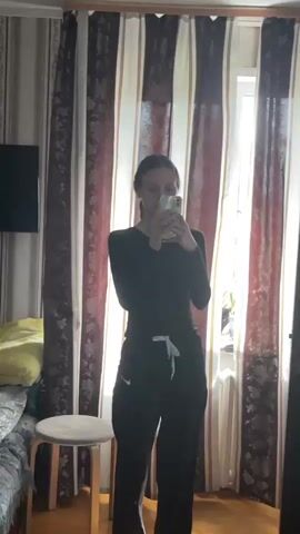The video shows two indoor scenes.

The first scene (0:00-0:01) features a young woman standing in front of a mirror, taking a selfie with her smartphone. She is wearing a black long-sleeved top and black sweatpants with a white drawstring and a Nike logo. The room has striped curtains (red, brown, and white), a window letting in daylight, a bed with a yellow pillow visible, a small white stool, and a television mounted on the wall.

The camera then quickly pans (0:01-0:02), showing a blurry view of curtains and what appears to be shelves.

The third scene (0:02-0:03) shows a man walking into a different room, carrying a white and gray vacuum cleaner with its hose attached. He is wearing a light-colored t-shirt and dark shorts. This room is also brightly lit by a window covered with sheer curtains. A bed with blue bedding, a bedside table with a lamp, and several balloons (light pink/beige) are visible. A banner with "HAPPY BIRTHDAY" written on it can be seen on the wall in the background. The overall setting is a home in Ulukulevo, Russia, during the daytime.
