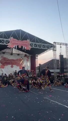 The video shows an outdoor performance of a traditional dance. Several male dancers, dressed in colorful patterned skirts and dark tops, are on a black paved area in front of a stage. Some dancers are on their knees, while others are in more dynamic poses, suggesting coordinated movements. Pink flower petals are scattered on the ground around the dancers. A stage with a banner reading "DHARMA NUSAWARA" is visible in the background, along with an audience seated on chairs and on the ground. The lighting suggests it is late afternoon or early evening. The overall environment appears to be a public gathering or festival.