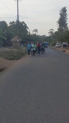 A group of approximately a dozen young men are jogging along a paved road in a rural area. They are dressed in a variety of casual athletic wear, including t-shirts, shorts, and running shoes, with some wearing soccer jerseys. A light blue passenger van follows the group from a distance. The road has a dirt shoulder on one side, with a small, wooden structure visible in the background. Power lines run above the road. The weather is overcast, suggesting either early morning or late afternoon.