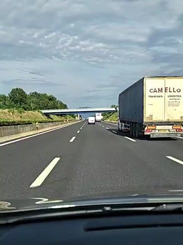 This video shows a view from inside a vehicle driving on a highway in Cassino, Italy. The highway has multiple lanes and is flanked by green grassy slopes and trees. A large white truck with "CAM TRASPORTI" written on its side is prominently featured on the right side of the road, driving in the same direction as the camera. Other vehicles, including cars and another truck, are visible further ahead on the highway. A pedestrian bridge spans across the highway in the distance. The sky is cloudy, suggesting it might be overcast or late afternoon. The overall scene depicts a typical highway journey.