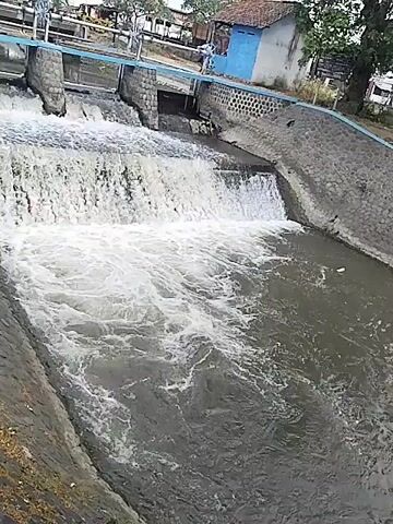 The video shows a waterfall from a man-made dam. Water flows over a concrete structure, creating a white, foamy cascade into a darker body of water below. The sides of the channel are lined with stone walls. In the background, there is a small blue building with a red-tiled roof, and some vegetation, including trees and bushes. There are a few people visible in the background, standing near the top of the dam. The overall scene depicts an operational water management structure in a rural or semi-urban setting.