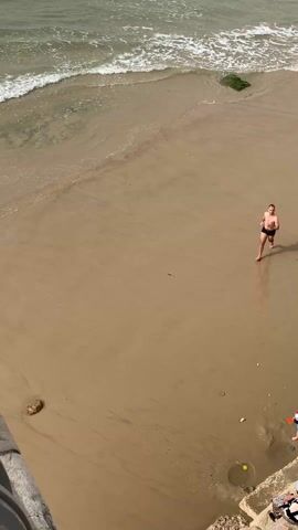 A high-angle shot depicts a sandy beach where two individuals, an adult male in black swim trunks and a younger male in a dark t-shirt and shorts, are running barefoot toward the camera. The adult male leads, with the younger one trailing slightly behind. The beach is wet, and the shoreline is visible with gentle waves breaking. At the lower right corner of the frame, there is a small child playing near a retaining wall. A ball rests on the ground. The scene is well-lit, suggesting it is daytime.