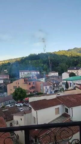 The video shows a view of a town nestled in a valley with hills covered in trees in the background. The sky is clear and blue, with some scattered clouds. In the foreground, there is a balcony with a black metal railing. The town consists of houses with red tile roofs and buildings in various colors, including pink, orange, and white. There are cars parked on a street visible in the middle ground. Above the town, there is a plume of dark smoke rising into the sky, originating from the direction of the wooded hills. Several smaller puffs of smoke and what appear to be small explosions or bursts of light are also visible in the sky, suggesting fireworks or possibly a small explosion. The overall scene depicts a daytime view of a town where an event involving smoke and possibly fireworks is occurring.