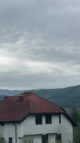 The video depicts a landscape observed from a moving vehicle. The sky is overcast with gray clouds. Initially, a glimpse of a white house with a red-tiled roof and a brick chimney is visible behind some green trees. The view then shifts to reveal a panorama of rolling green hills and valleys, covered with trees. As the vehicle progresses, the camera's motion blurs the foreground, showcasing only streaks of green foliage. The occasional blurred image of buildings reappears intermittently through the trees.