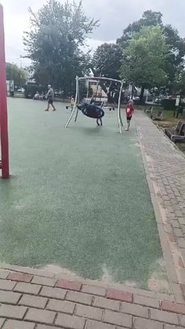 A man is sitting on a blue tire swing at a playground. He is wearing a blue shirt and jeans and sunglasses. A young girl is standing next to the swing set, holding onto one of the poles. Another child, a girl with blonde hair, is on the ground near the swing set. In the background, there are trees and buildings. The ground is covered in a green artificial turf.