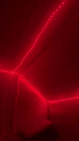 The video shows a room illuminated by red LED strip lights. The lights are installed along the ceiling and walls, creating a vibrant red ambiance. The walls and ceiling appear to be made of wood planks. In the lower right portion of the frame, a dark object is visible, possibly a piece of furniture or a bed. The room is dimly lit except for the red lights, which cast a strong glow on the wooden surfaces. There are no people visible in the video. The camera pans slowly, capturing the way the lights wrap around the corners of the room and highlight the texture of the wood.