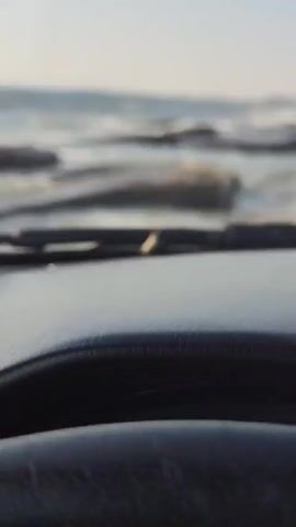 The video shows a close-up view from inside a vehicle, looking out through the windshield. A small, light-colored insect, possibly a grasshopper, is visible on the dashboard in the foreground. Beyond the windshield, the ocean is visible, with waves crashing against a rocky shoreline. The water appears choppy and there are large, flat rocks partially submerged in the water. In the distance, a structure resembling a crane or tower can be seen near the coast. The sky is clear and the overall lighting suggests it is daytime. The vehicle appears to be moving slowly or stopped near the water's edge.