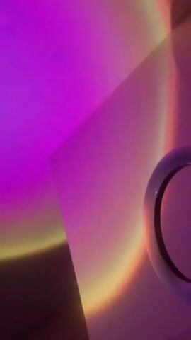 The video shows a selfie ring light on a stand, casting a purplish pink light with a golden yellow edge onto a wall and ceiling. The camera moves slightly, showing the ring light and then focusing on the diffused light on the wall. There's also a glimpse of a white bed or surface beneath the ring light.