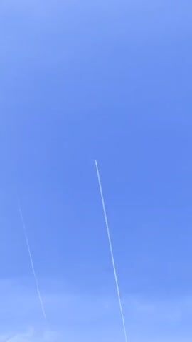 The video shows a clear blue sky with two white contrails left by airplanes. The contrails are thin lines stretching diagonally across the frame, indicating the planes are flying at a high altitude. The angle of the contrails suggests the planes are moving upwards or away from the viewer. The sky is a consistent shade of blue, with no clouds visible. The camera appears to be stationary, focusing on the contrails as they persist in the sky.
