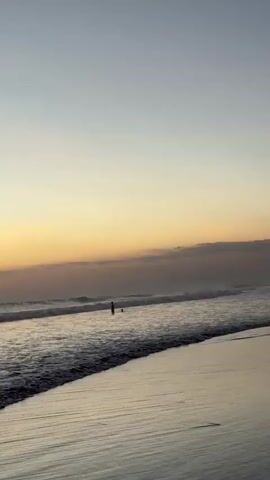 A person stands in the shallow ocean water on a beach during what appears to be sunset or sunrise. The sky is a gradient of pale blue and soft orange, with some clouds visible. The ocean is calm with small waves breaking near the shore. The beach is wet and reflective, with the gentle slope of the sand visible in the foreground. The person is silhouetted against the water and sky, making their features indistinct. The overall atmosphere is tranquil and serene.