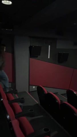 The video captures a young woman dancing and moving playfully in an empty movie theater. She has long dark hair and is wearing a dark long-sleeved top, blue jeans, and white sneakers. The theater is dimly lit, with rows of red and black seats visible in the foreground and along the sides. Red panels decorate the lower sections of the walls.

At the beginning of the video, she is seen moving from the left side of the frame, walking down an aisle between seat rows, with her arms slightly outstretched. As she continues to move towards the right and closer to the front of the theater, a large projection screen becomes visible. The screen is displaying white end credits against a dark background. She then turns to face the camera, smiling broadly, and continues her dance-like movements with her arms, occasionally making a small twirl. The ambiance is dark, with light primarily coming from the projector screen and some overhead ceiling lights. The woman appears happy and energetic throughout the short clip, enjoying her impromptu dance in the empty cinema. Upbeat music is playing in the background.