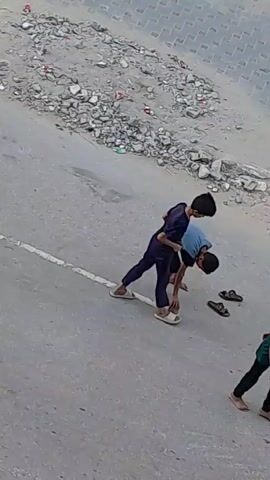 Two boys are playing on a street. One boy is wearing a dark blue outfit, and the other is wearing a light blue t-shirt and dark shorts. Their shoes are on the ground nearby. They appear to be engaged in a playful interaction, possibly a game of tag or a mock fight. The environment is a paved street with debris scattered on the side, suggesting an urban or semi-urban setting.