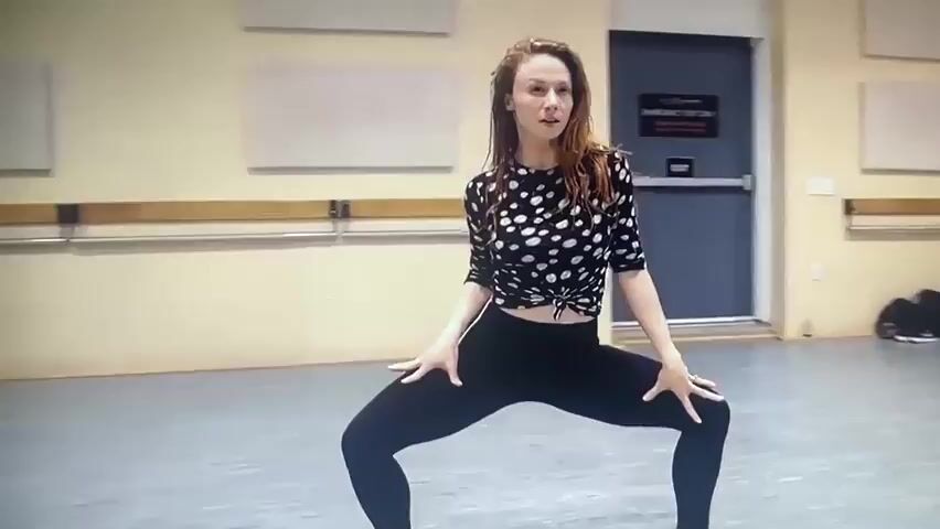 A woman with long, auburn hair is dancing in a dance studio. She is wearing a black, long-sleeved shirt with white polka dots, black leggings, and black heeled boots. She is in a wide-legged squat with her knees bent and her hands resting on her knees. The studio has a grey floor, beige walls, and wooden barre. There is a door in the background with a sign that says "EMERGENCY EXIT ONLY." There are also several bags and shoes in the background. The woman moves her arms and torso in a fluid motion, then extends one leg forward and her arms upward, before bringing her hands to her stomach and bending backward.