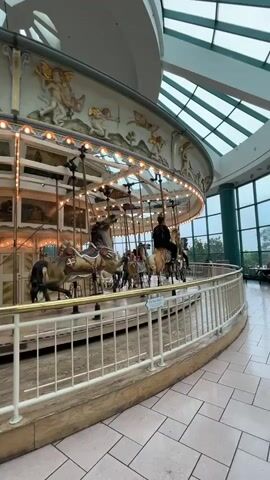 The video pans across a carousel situated indoors, likely in a mall or similar public space, indicated by the tiled flooring and large windows with a green vertical support column visible. The carousel is ornately decorated with cream and gold accents, featuring various horse figures. Several horses are visible, some with riders, including one person wearing dark clothing. The carousel is illuminated by string lights along its upper edge. Above, a modern skylight with a visible grid structure allows natural light into the space. The overall atmosphere appears calm and perhaps slightly overcast due to the lighting conditions visible through the windows.