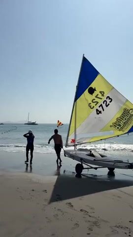 Two people are on a sandy beach with a small sailboat on a trailer. The sailboat has a yellow, blue, and white sail with the number 4723 printed on it. The person on the left is wearing dark clothing and a hat. The person on the right is wearing dark shorts and is shirtless. They are both facing the water, and the sailboat is partially in the water. The sun is shining, and the sky is clear. In the background, there are other boats and a pier. The waves are small, and the water is calm. The people appear to be preparing to launch the sailboat into the water.