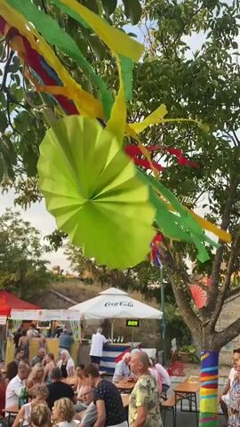 This is a short video taken at an outdoor event. Colorful streamers and paper fans are hanging from trees in the foreground, decorated with vibrant green, yellow, blue, and red colors. In the background, a crowd of people is gathered, seated at tables and standing around. There are several stalls with tents and umbrellas, one of which has a Coca-Cola logo. People appear to be socializing and enjoying themselves. The overall atmosphere seems festive and lively.