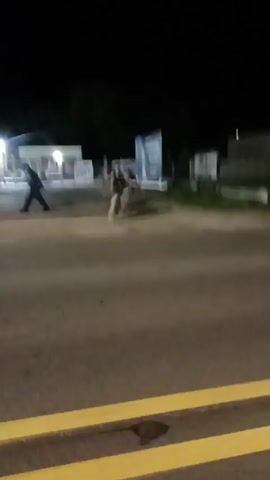At night, a video shows three people standing on a dark street. In the background, there are some businesses and billboards. One of the people picks up a garbage can and lifts it up. He then drops it on the ground.