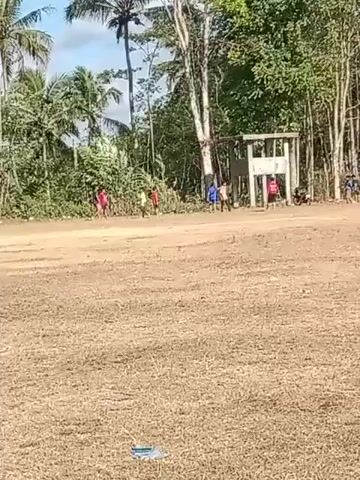 The video shows a group of children playing in a dirt field with trees and vegetation in the background. There is a partially constructed building or structure on the right side of the frame. The children are scattered across the field, some appearing to be running or playing a game. The ground is dry and dusty. The sky is visible and appears to be blue with some clouds.