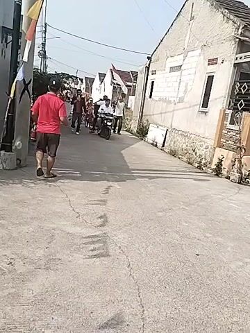 A person in a red shirt walks away from the camera down a narrow, paved street. They are on the left side of the street, next to a utility pole decorated with red, white, and yellow flags. In the distance, a group of people is walking towards the camera. Some are carrying Indonesian flags. One person is riding a motorcycle and another is riding a bicycle. Houses with white walls and tiled roofs line both sides of the street. The sky is clear and bright.