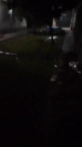 The video shows an outdoor scene at night. It is dark, and the camera is shaky, suggesting it may be handheld and possibly in motion. There is evidence of rain or water on the ground, with reflections visible under what appears to be artificial light. In some frames, a well-lit doorway or porch is visible, with a light fixture casting a bright glow. There are also indistinct shapes that could be trees or other foliage, and possibly parts of buildings. The overall impression is of a dark, wet, outdoor environment at night. There are no clearly identifiable people or specific objects in focus.