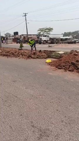 The video shows a man in a yellow safety vest standing on a pile of reddish-brown dirt in the middle of an asphalt road. He appears to be surveying the area. A shovel is visible near him. Power lines run overhead. Across the road, there are various buildings and vehicles, including a large white truck. Several people are visible in the background on the side of the road near some stalls. The sky is overcast.