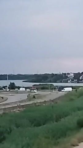 The video displays a body of water in the background with a shoreline characterized by trees and buildings. In the mid-ground, a road is visible with several vehicles, including cars and trucks, driving on it. The foreground consists of grassy terrain. The sky appears overcast. The camera appears to be moving from left to right, capturing a panoramic view of the landscape.