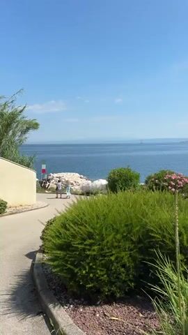This is a vertical video filmed outdoors on a sunny day, likely in the late morning or early afternoon. The scene is a pathway leading to a beautiful coastal view of the Adriatic Sea. The sea is calm and a deep blue, with a few distant islands or landmasses visible on the horizon. The sky is a clear, bright blue with a few wispy white clouds.

In the foreground, the pathway is paved and curves to the right. It's bordered by lush, well-maintained greenery, including manicured bushes and flowering plants with pink blooms. Some of these plants are in raised garden beds filled with white and dark pebbles. Further down the path, there are more trees and shrubs.

On the left side of the pathway, a light-colored building is partially visible. Adjacent to it, a small Italian flag is mounted on a pole, suggesting a connection to Italy or a preference for Italian aesthetics.

As the pathway approaches the sea, it opens up to a scenic overlook. Here, two people, appearing to be women, are walking and looking out at the water. They are dressed casually in light-colored clothing. Beyond them, there is a rocky shoreline leading down to the sea.

Further along the coast, several white beach umbrellas are set up on what appears to be a beach or a lounging area near the water. Some white structures, possibly sun loungers, are visible under the umbrellas. The overall atmosphere is serene and idyllic, suggesting a peaceful vacation or leisure setting. The clear weather and bright sunshine contribute to a feeling of warmth and tranquility.