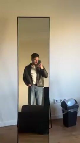 A man stands in front of a full-length mirror, holding a phone up to take a picture of himself. He is wearing a white t-shirt, blue jeans, and a dark jacket. The mirror has a thin black frame. The room has light-colored walls and a wooden floor. Behind the man and to the side, there is a dark colored couch or chair and a waste bin with a white lid. The lighting is bright.