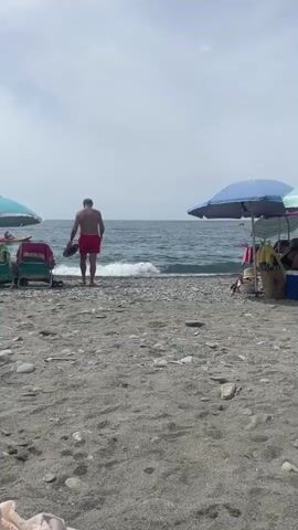 A person wearing red swim trunks walks towards the ocean on a pebble beach, with umbrellas and lounge chairs set up nearby. The person stops at the edge of the water and appears to be taking something from their face. Waves crash gently on the shore under a cloudy sky.