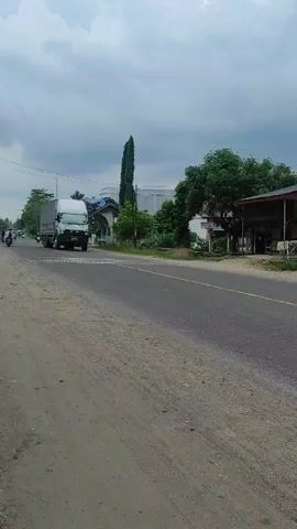 The video captures a daytime scene on a two-lane asphalt road in Lancang Barat, Indonesia, under an overcast sky. A large white Isuzu Giga truck with "ISUZU GIGA" and "S1100" visible on its cab is seen traveling from left to right. The truck has a covered cargo area.

In the foreground, a scooter with two riders passes, also moving from left to right. Another scooter and a motorcycle can be seen further down the road. The road has a wide, unpaved dirt shoulder on the left. Along the right side of the road, there are several green trees, including a tall, slender tree, and some modest buildings or shops. Power lines run along the left side of the road. The overall impression is of a semi-rural or outer urban area with moderate traffic.