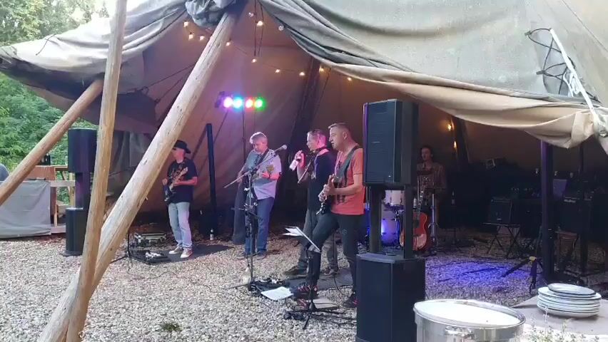 A band is playing music under a large tent. There are five men visible on stage, playing instruments and singing. The band appears to be a rock band, with electric guitars and drums. The tent is decorated with string lights, and there are speakers set up on the stage. The ground is covered in gravel. A person is partially visible on the left side of the screen, sitting at a table. There are trees and foliage visible behind the tent. The band members are dressed in casual clothing. The lighting is dim, with colorful stage lights illuminating the performers.