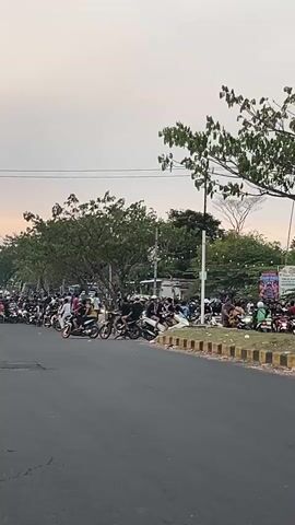 A wide shot shows a street scene with many people and motorcycles. The sky is overcast with a hint of sunset colors. On the left side of the street, a line of motorcycles and people are gathered. Some individuals are seated on motorcycles, while others stand around. Further down the street, more people and motorcycles are visible, forming a crowd. The right side of the street has a sidewalk with more people sitting and standing, and parked motorcycles. A row of trees runs along the side of the street, and a few utility poles are visible in the background. The road appears to be asphalt, and the weather seems mild. The overall impression is of a social gathering or an event involving motorcycles.