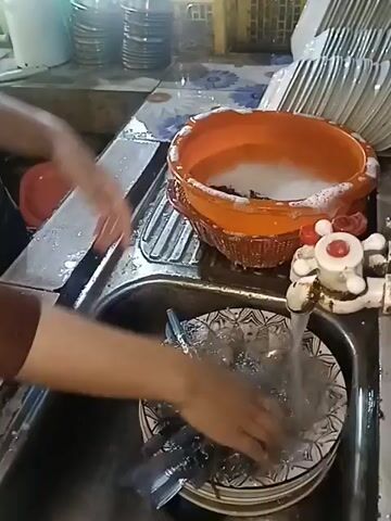 A pair of hands, one wearing a brown sleeve, is seen washing kitchen utensils under a running faucet. The utensils, appearing to be cutlery and plates, are in a stainless steel sink. To the right of the sink is an orange basin filled with soapy water. In the background, shelves hold stacked dishes and other kitchen items. The scene appears to be indoors, likely a kitchen or food preparation area.