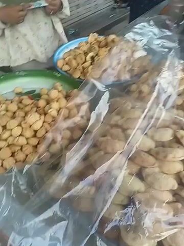The video shows a street-side stall selling snacks. The stall is covered by a makeshift roof. In the foreground, there are several clear plastic containers filled with various snacks. Some snacks are small and golden brown, while others are larger and crescent-shaped. Behind these, there are stacks of packaged snacks, some appearing to be cookies or crackers. A woman wearing a blue hijab is standing behind the counter, and another person, possibly a customer, is interacting with the items on display. A motorcycle is parked to the right of the stall. A large banner hangs from the stall, displaying the name "DAPUE BHOI KAKLA" and showing images of snacks. The background reveals a road with a few cars passing by and lush greenery. The overall scene suggests a small business operating outdoors.