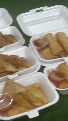 The video shows a food stall or outdoor dining area. In the foreground, there are several white styrofoam containers holding spring rolls with a dipping sauce and what appears to be fried chicken. To the right, a large tray is filled with numerous pieces of fried chicken. The surfaces are covered with a green material, possibly artificial turf. In the background, there are stacks of white containers and some blurred figures of people. The lighting suggests it might be evening or an indoor setting with artificial light. The camera is in motion, giving a slightly blurry and unstable view of the scene.