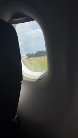 This video captures a view from an airplane window as the aircraft taxis on the tarmac. The camera is positioned to the left of the window, with a dark airplane seat in the foreground obscuring part of the view. The window itself is oval-shaped and reveals a bright outdoor scene. Outside, the ground appears to be a runway or airport area with green grass and paved surfaces. Several buildings, possibly airport terminals or hangars, are visible in the distance, characterized by light-colored walls and blue roofs. The sky is partly cloudy with a mix of white clouds and patches of blue. The airplane is moving, as evidenced by the slight blur and shifting perspective of the landscape outside the window. The overall impression is one of preparing for takeoff or having just landed.