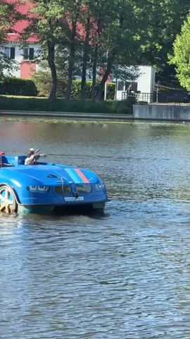 A blue pedal boat designed to resemble a convertible car moves across a body of water. The boat has red, white, and blue racing stripes on its hood and sides. In the boat, a young person wearing a red cap and a child wearing a red and white striped shirt and a red cap are seated. Another child, wearing a white cap, is seated in the front of the boat and appears to be steering. The water is calm with slight ripples. In the background, there is a grassy area with trees and a red-roofed building. The lighting suggests it is daytime with sunshine.