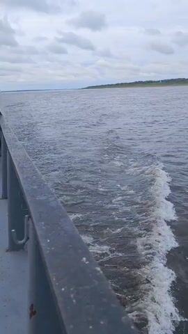 The video shows a view from a boat moving through water. The boat's railing is visible in the foreground, taking up the left side of the frame. Behind the railing, the water is disturbed, creating a wake with white foam and churning waves that trail behind the boat. The water extends towards a distant horizon line where the sea meets a cloudy sky. The overall impression is one of forward motion on a body of water.